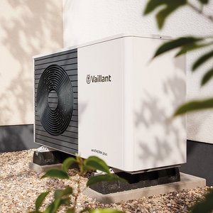 Eine moderne Wärmepumpe steht auf einem Kiesboden neben einem hellen Wohnhaus, umgeben von Pflanzen und einem gemütlichen Sitzbereich.