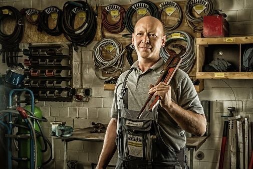 Bernd Heiker in Arbeitskleidung der eine große Wasserpumpenzange auf der der Schulter trägt